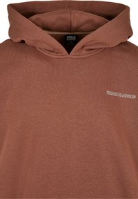 Sudadera de algodón marrón con un bolsillo tipo canguro en el frente, capucha con cordón y logo bordado en blanco en el lado izquierdo del pecho.