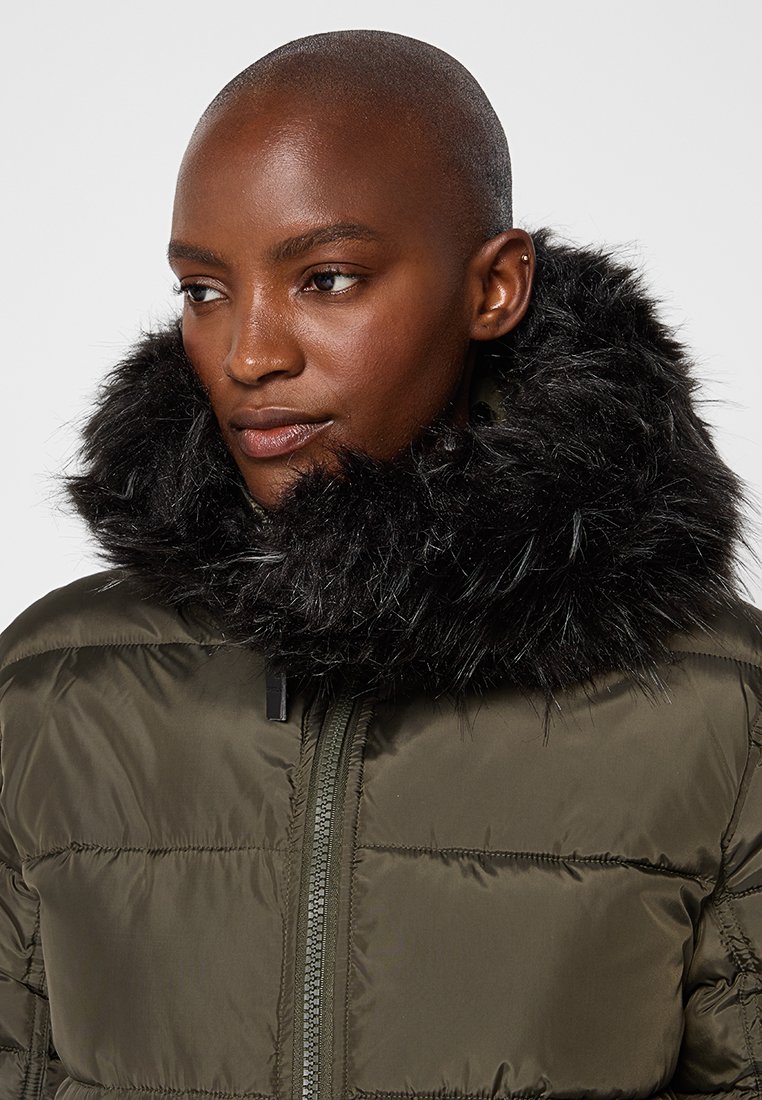 Persona con cabeza rapada que lleva una chaqueta acolchada verde oscura con capucha de falso pelo negro, mirando ligeramente hacia un lado.