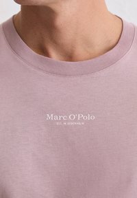 Zbliżenie osoby w jasnofioletowej koszuli z białym nadrukiem "Marc O'Polo EST. W Sztokholmie" na piersi.