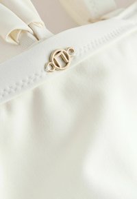 Tissu blanc à texture lisse, doté d'une boucle circulaire décorative dorée et d'une couture le long du bord.