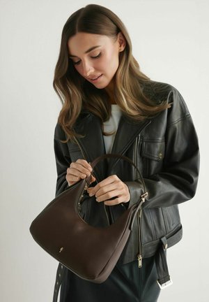 Kobieta w czarnej skórzanej kurtce trzymająca i rozpinająca ciemnobrązową skórzaną torebkę na jednolitym tle.
