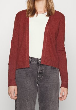 Woman wearing a rust red open-front cardigan over a white top and high-waisted faded black jeans against a plain background.