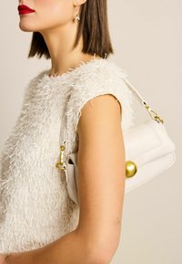 Sac en cuir blanc avec un accent doré arrondi, tenu par une personne portant un haut blanc texturé. Design lisse et structuré.