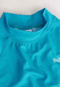 Türkiissinise varjukaitsega kõrge kaelusega, siledast venivast kangast, flatlock-õmblustega ja valge logoga alumisel paremal küljel.