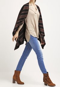 Sciarpa a righe nera e borgogna, design aperto, indossata sopra un top beige. Jeans skinny blu e stivali alla caviglia marroni con frange.