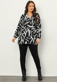 Schwarz-weiß gemusterte Tunika mit V-Ausschnitt und langen Ärmeln, kombiniert mit schmalen schwarzen Hosen und schwarzen Absatzstiefeln. Glatte Stofftextur.