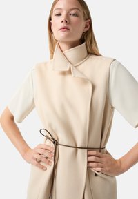 Beige ärmelloser Mantel mit hohem Kragen, strukturiertem Stoff und tailliertem Schnitt; kombiniert mit kurzen weißen Ärmeln. Minimalistisches Design, praktische Taschen.