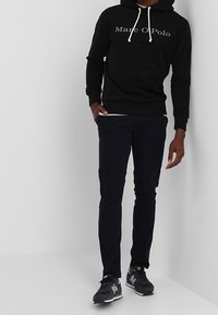 Muž v černé mikině Marc O'Polo, tmavých slim kalhotách a černých teniskách New Balance stojí proti bílé rohové zdi.