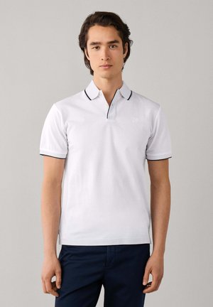 Jonge man draagt een wit poloshirt met zwarte bies en donkerblauwe broek, staand tegen een effen lichte achtergrond.