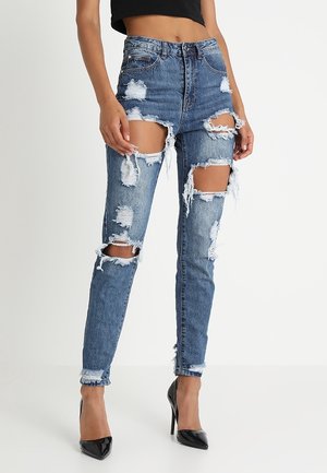 Femme portant un jean bleu déchiré taille haute avec de grandes déchirures, associé à des talons hauts noirs et un top court noir, sur un fond uni.