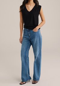 Kvinna i ärmlös svart V-ringad topp, lösa blåjeans och svarta öppna klackskor, med ena handen i fickan, stående framför en enfärgad bakgrund.