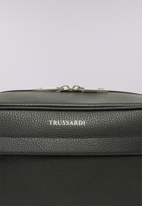 Černá texturovaná kožená taška se stříbrným dvojitým zipem a bílým logem "TRUSSARDI" umístěným uprostřed pod zipem na světlém jednobarevném pozadí.