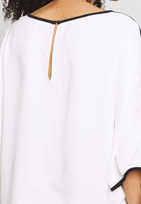 Blouse blanche avec des manches courtes évasées, un accent noir et une ouverture en forme de clé dans le dos, en tissu léger et lisse.