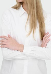 Femme aux longs cheveux blonds portant une chemise blanche boutonnée, les bras croisés sur la poitrine, légèrement tournée vers la gauche.