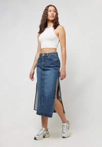 Femeie purtând un top scurt alb, fustă midi din denim cu talie înaltă și tăieturi laterale, și pantofi sport Nike albi, stând pe un fundal gri simplu.