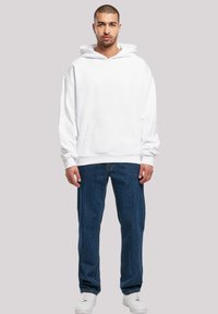 Hvit hettegenser med kengurulomme, avslappet passform og snor i hetten, kombinert med blå denimjeans og hvite joggesko.