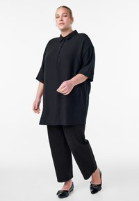 Schwarze Tunika mit einem kragenlosen Ausschnitt, kurzen Ärmeln und zwei vorderen Taschen, kombiniert mit weit geschnittenen schwarzen Hosen und flachen schwarzen Schuhen.