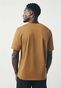 Brun bomulds t-shirt med korte ærmer og rund halsudskæring. Glat tekstur og afslappet pasform, set fra ryggen mod en lys baggrund.