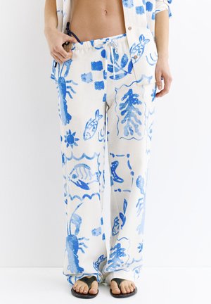 Femme portant un pantalon blanc ample et une chemise assortie avec des imprimés bleus représentant la vie marine, tenant la ceinture, pieds nus avec des sandales noires à doigts.