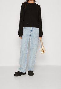 Suéter negro de punto con diseño texturizado, jeans de mezclilla azul claro, mocasines gruesos negros y un pequeño bolso de mano dorado.