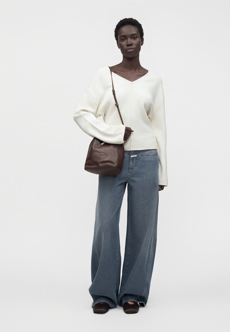 Femme portant un pull blanc à col en V, un jean bleu à jambes larges, des chaussures marron et un sac à épaule marron sur un fond uni.
