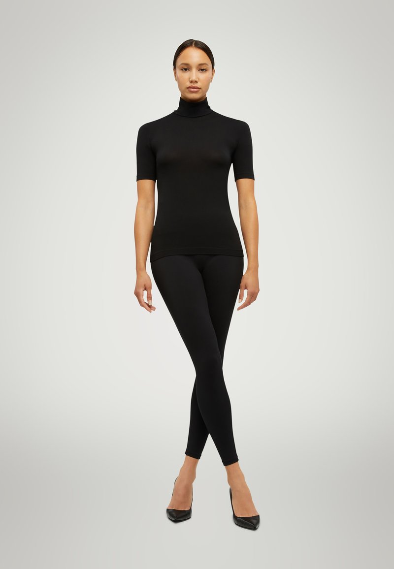 Maglione aderente nero a collo alto con maniche corte, abbinato a leggings neri e décolleté neri a punta. Texture liscia e design senza cuciture.