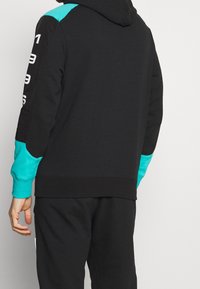 Schwarzer Kapuzenpullover mit türkisfarbenen Akzenten an Schultern und Bündchen sowie weißen Textdetails an den Ärmeln. Weicher Stoff.