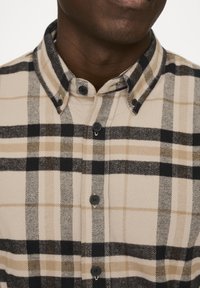 Beige kariertes Flanellhemd mit schwarz und tanfarbener Karomuster, ausgestattet mit einem Button-Down-Kragen und schwarzen Knöpfen entlang der Vorderseite.