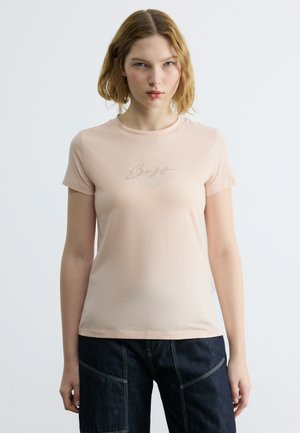 Νεαρή γυναίκα με ξανθά μαλλιά μέχρι τους ώμους, που φοράει ένα ανοιχτό ροζ εφαρμοστό μπλουζάκι με γραπτό κείμενο και σκούρο παντελόνι φαρδιάς γραμμής.