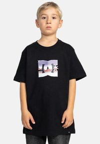 Černé bavlněné tričko s barevným designem loga DC. Krátké rukávy, kulatý výstřih, volný střih. Jednoduchá textilie a čisté linie.