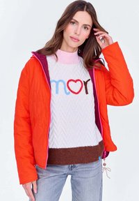 Kobieta z długimi brązowymi włosami, ubrana w pomarańczową pikowaną kurtkę na białym swetrze z napisem "amor" i niebieskie dżinsy, stojąca na białym tle.
