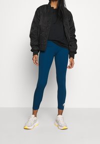 Svart bomberjacka, svart t-shirt, teal leggings med vitt logotyp, och grå sneakers med gula accenter. Slät textur och skräddarsydd design.