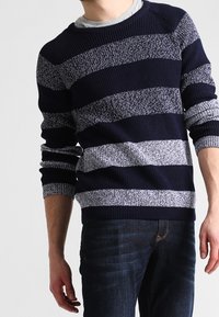 Homme portant un pull en maille rayé bleu marine et gris et un jean bleu foncé, debout devant un fond uni.