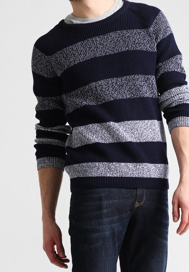 Homme portant un pull en maille rayé bleu marine et gris et un jean bleu foncé, debout devant un fond uni.