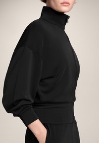 Černá mikina na zip s vysokým límcem, objemnými rukávy a přiléhavým pasem. Vyrobena z měkké, texturované tkaniny.