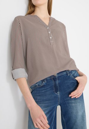 Femme portant un chemisier à rayures verticales marron et blanches, boutonné, avec les manches retroussées, et un jean en denim bleu, posant devant un fond blanc.