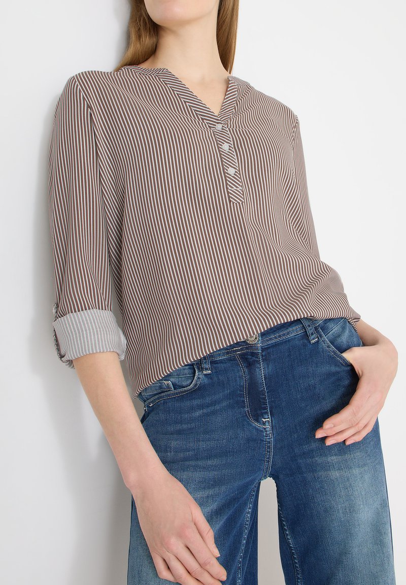 Femme portant un chemisier à rayures verticales marron et blanches, boutonné, avec les manches retroussées, et un jean en denim bleu, posant devant un fond blanc.