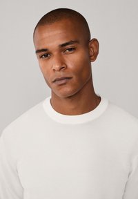 Maglione bianco a maglia con scollo rotondo, caratterizzato da un tessuto textured. Il modello ha i capelli corti e un'espressione neutra su uno sfondo semplice.