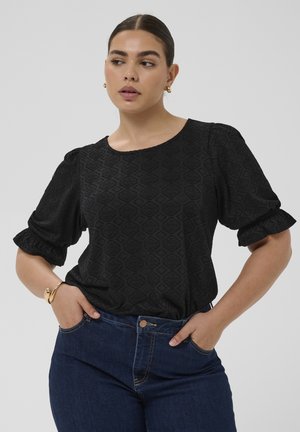 Femme aux cheveux foncés attachés, portant un haut noir texturé à manches bouffantes et un jean bleu, mains dans les poches, regardant sur le côté.