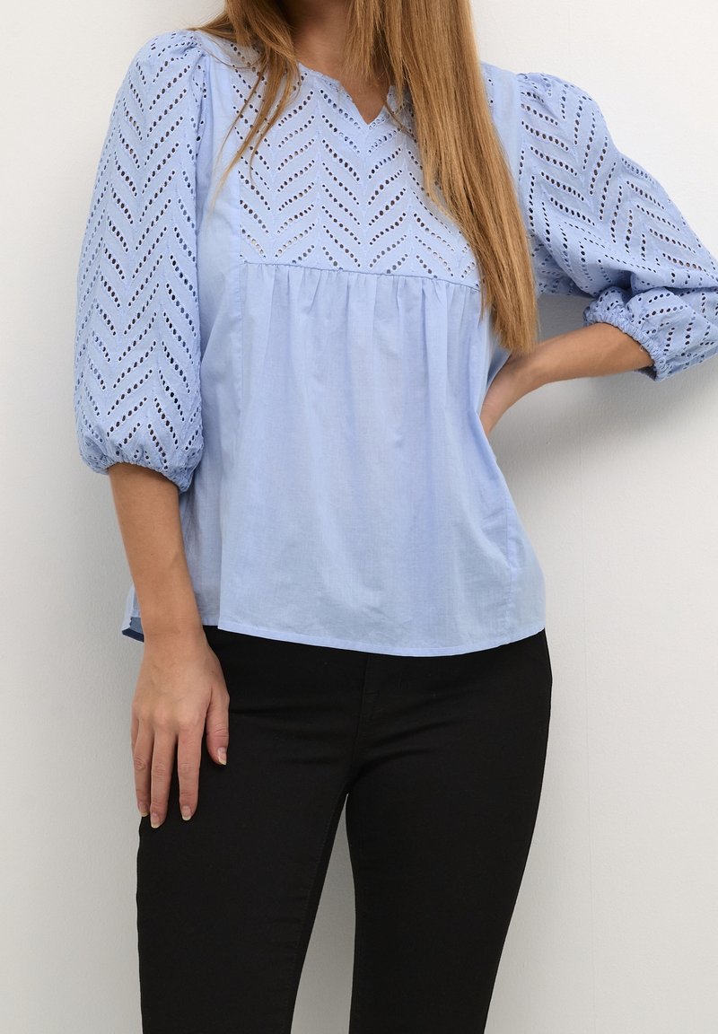 Blouse bleu clair avec un col en V, présentant un motif découpé en chevrons texturé sur les manches et le corps, associée à un pantalon noir ajusté.