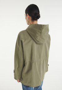 Oliwkowa kurtka z kapturem o luźnym kroju, wyposażona w ściągacz w talii i guzikowe mankiety, wykonana z trwałej tkaniny.