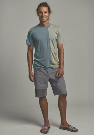 Mann mit lockigem Haar in einem blau-beigen T-Shirt mit Farbteilung, grauen Shorts und gestreiften Slide-Sandalen, der vor einem einfarbigen Hintergrund steht und lächelt.