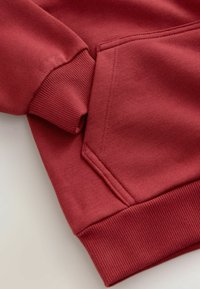 Nærbillede af en rød sweatshirtærme, ribbet manchet, kengurulomme og ribbet talje på en lys baggrund.