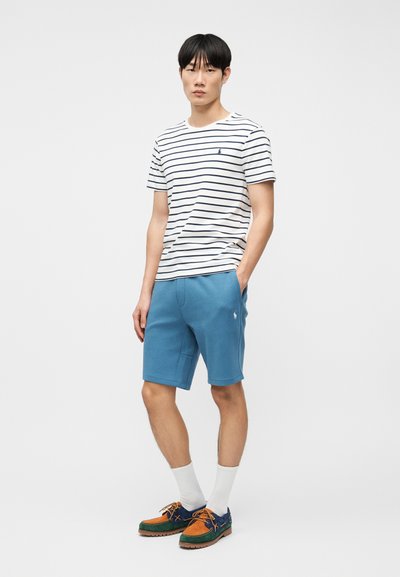 Νεαρός άνδρας στέκεται με τα χέρια στις τσέπες, φορώντας λευκό και ναυτικό ριγέ μπλουζάκι, μπλε σορτς, λευκές κάλτσες και πολύχρωμα αθλητικά παπούτσια.