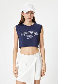 Granatowy, bezrękawnikowy crop top z napisem "LOS ANGELES 1992 CALIFORNIA" w białym kolorze; zestawiony z białą spódnicą w kroju kopertowym i białą czapką.