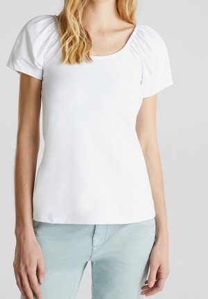 Kobieta w białej bluzce z krótkim rękawem z marszczonymi detalami na ramionach i jasnoniebieskich spodniach, stojąca na jednolitym tle.