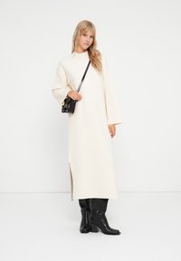 Robe longue couleur crème avec des manches larges, une fente sur le côté et un décolleté simple ; associée à des bottes en cuir noires jusqu'aux genoux et à un petit sac noir.
