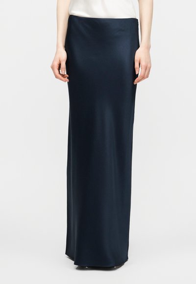 Femme portant une jupe longue en satin bleu marine associée à un haut blanc, debout devant un fond blanc uni.