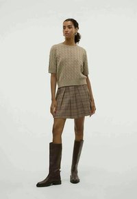 Pull en maille torsadée beige à manches courtes associé à une jupe plissée à carreaux marron et des bottes en cuir marron jusqu'aux genoux. Boucles d'oreilles circulaires simples.