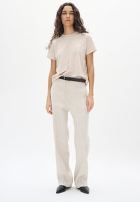 Beige T-shirt met korte mouwen, gecombineerd met lichtgekleurde linnen broeken, een zwarte riem en puntige zwarte schoenen, tegen een effen witte achtergrond.